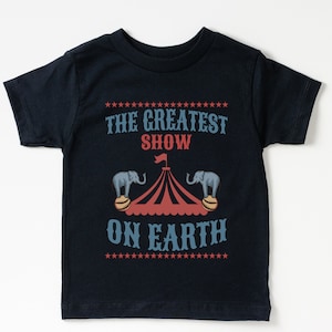 May include: A black t-shirt with a white and red circus tent graphic and the text "The Greatest Show On Earth" printed on it.