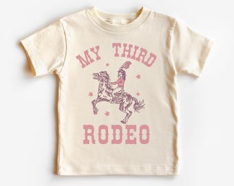 May include: A cream colored t-shirt with a pink graphic of a girl riding a horse. The graphic says "My Third Rodeo" with stars around the graphic.