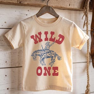 May include: A cream-colored short-sleeve t-shirt featuring a red graphic print that reads WILD ONE. Between the words is a blue illustration of a cowboy riding a bucking horse. The design has a distressed texture and is centered on the front.