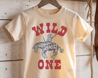 May include: A cream-coloured short-sleeve t-shirt featuring a red graphic print that reads WILD ONE. Between the words is a blue illustration of a cowboy riding a bucking horse. The design has a distressed texture and is centred on the front.