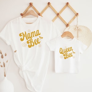May include: Two white t-shirts with a bee theme. The larger shirt says "Mama Bee" in a retro font with a bee and honeycomb design. The smaller shirt says "Queen Bee" in the same font with a bee and honeycomb design.