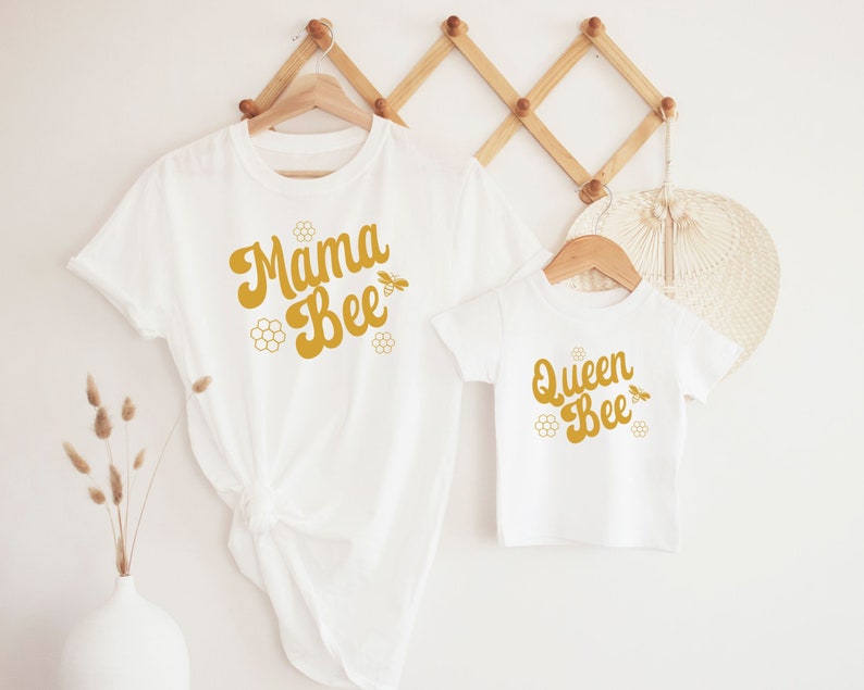May include: Two white t-shirts with a bee theme. The larger shirt says "Mama Bee" in a retro font with a bee and honeycomb design. The smaller shirt says "Queen Bee" in the same font with a bee and honeycomb design.