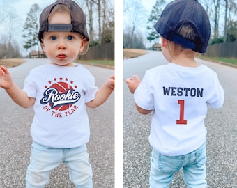 May include: A white t-shirt with a basketball graphic and the text "Rookie of the Year" on the front. The back of the shirt has the name "WESTON" and the number "1" in red.