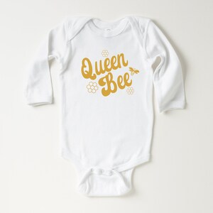 May include: White long-sleeve baby onesie with a gold graphic that says "Queen Bee" with a bee and honeycomb design.