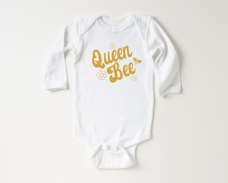 May include: White long-sleeve baby onesie with a gold graphic that says "Queen Bee" with a bee and honeycomb design.