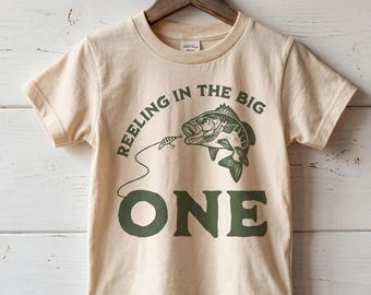 以下が含まれることがあります： ベージュの半袖Tシャツで、釣り糸にかかった魚のダークグリーンのグラフィックと、ダークグリーンの文字で「REELING IN THE BIG ONE」とプリントされています。Tシャツは白い木製の背景の前で木製のハンガーに掛けられています。