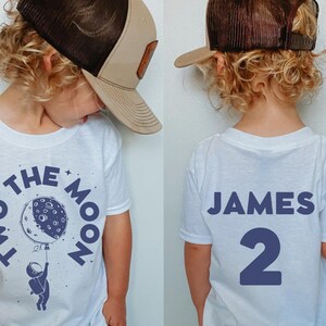 May include: White t-shirt with a blue astronaut holding a moon with the text "Two to the Moon" on the front. The back of the shirt has the name "James" and the number "2" in blue.