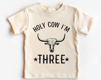 Könnte beinhalten: Ein hellbeiges T-Shirt mit einer schwarzen Grafik eines Bullenkopfes und dem Text "HOLY COW I'M THREE" mit Sternen auf beiden Seiten des Wortes "THREE".