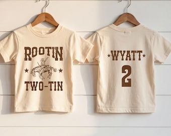 May include: Two cream-colored short-sleeve t-shirts hanging on wooden hangers. The left shirt features brown text reading ROOTIN TWO-TIN with a cowboy on a bucking horse. The right shirt features brown text reading WYATT and the number 2.