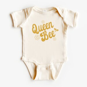 May include: A cream-colored baby onesie with a gold graphic that says "Queen Bee" with a bee and honeycomb design.