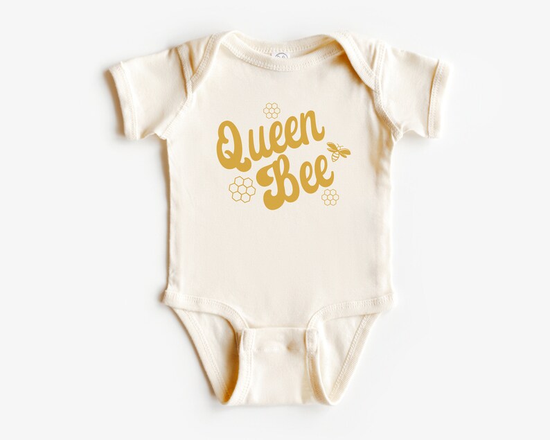 May include: A cream-colored baby onesie with a gold graphic that says "Queen Bee" with a bee and honeycomb design.