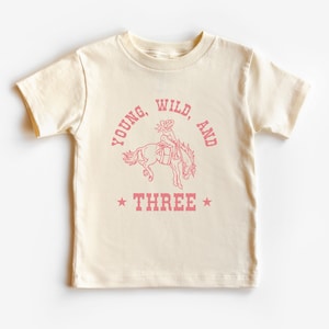 May include: A cream colored t-shirt with a pink graphic of a girl riding a horse. The text "Young, Wild, and Three" is printed on the shirt.