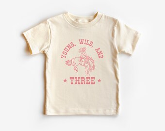 May include: A cream colored t-shirt with a pink graphic of a girl riding a horse. The text "Young, Wild, and Three" is printed on the shirt.