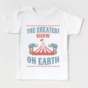May include: White t-shirt with a red and white circus tent graphic and two blue elephants balancing on balls. The text "THE GREATEST SHOW ON EARTH" is printed in blue on the shirt.