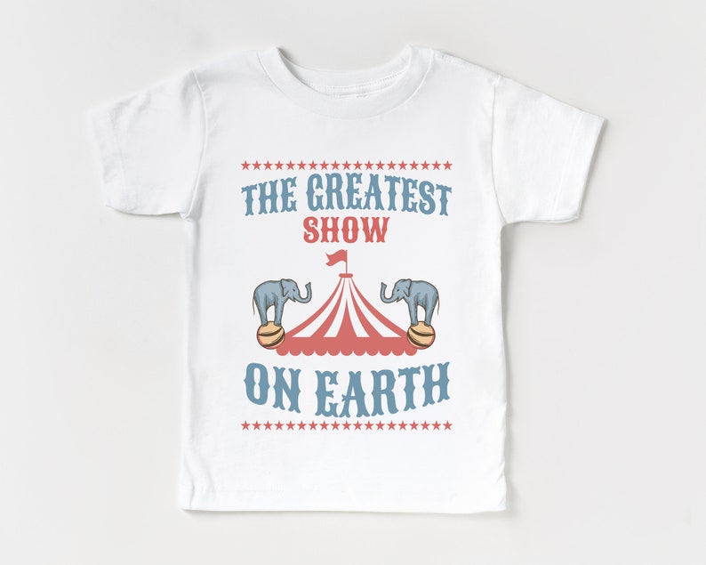 May include: White t-shirt with a red and white circus tent graphic and two blue elephants balancing on balls. The text "THE GREATEST SHOW ON EARTH" is printed in blue on the shirt.