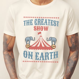 May include: White t-shirt with a vintage circus design. The text reads "The Greatest Show On Earth" with two elephants balancing on balls under a striped circus tent.