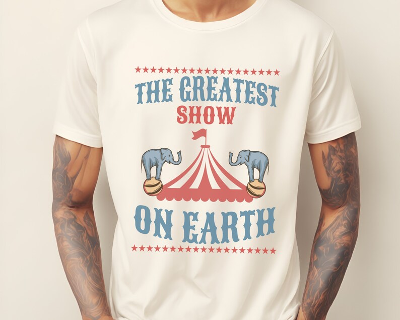 May include: White t-shirt with a vintage circus design. The text reads "The Greatest Show On Earth" with two elephants balancing on balls under a striped circus tent.