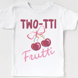 May include: White toddler t-shirt with the words "TWO-TTI Frutti" in burgundy and pink script. The design includes a pair of cherries with a pink bow.
