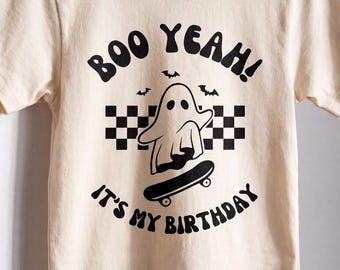 May include: A cream-colored short-sleeve t-shirt featuring a black graphic print of a ghost on a skateboard, a checkered pattern, small bats, and the text "BOO YEAH! IT'S MY BIRTHDAY" in a bold, rounded font centered on the front of the shirt.