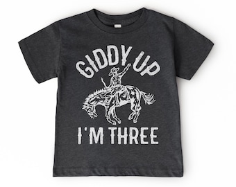May include: Dark gray t-shirt with a white graphic of a cowboy riding a bucking bronco. The text "GIDDY UP" is above the image, and "I'M THREE" is below. The shirt is made of a soft, comfortable material.