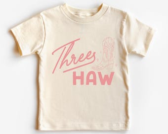 May include: Cream-colored t-shirt with the text "Threes Haw" in pink script and block letters. A pink cowboy boot graphic is also featured. This tee is perfect for a child's birthday or a western-themed event.
