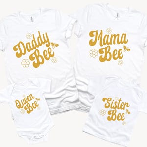 May include: A set of four white t-shirts with a bee theme. The shirts feature the text "Daddy Bee", "Mama Bee", "Queen Bee", and "Sister Bee" in a retro font with a bee illustration.