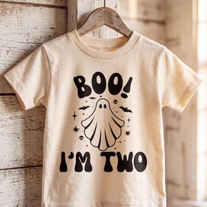 May include: A cream-colored short-sleeve t-shirt featuring a black graphic print. The design includes a central ghost illustration surrounded by small bats, spiders, stars, and skulls, with the text BOO! above and I'M TWO below the ghost.