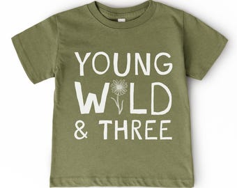 May include: Olive green t-shirt with the words "YOUNG WILD & THREE" in white block letters. A white daisy graphic is between the words "WILD" and "&". The shirt has short sleeves and a crew neck.