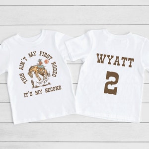 May include: White t-shirt with a brown graphic of a cowboy riding a bucking horse. The text on the shirt reads "This ain't my first rodeo, it's my second". The back of the shirt has the name "WYATT" and the number "2" in brown.