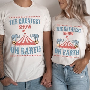 May include: Two white t-shirts with a circus theme. The shirts feature a red and white striped tent with blue elephants on either side. The text on the shirts reads "The Greatest Show On Earth".