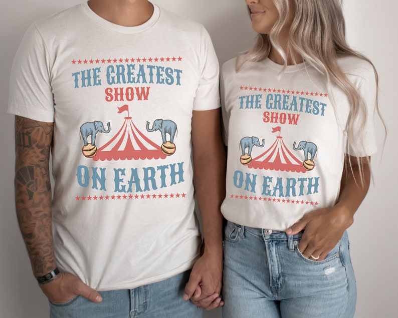 May include: Two white t-shirts with a circus theme. The shirts feature a red and white striped tent with blue elephants on either side. The text on the shirts reads "The Greatest Show On Earth".