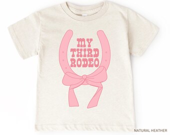May include: A light beige t-shirt with a pink horseshoe and bow design. The text "MY THIRD RODEO" is printed in pink on the shirt.