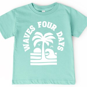 May include: A light teal t-shirt with a white graphic. The graphic features a palm tree, a setting sun, and a wave, with the words "WAVES FOUR DAYS" in a circular arrangement. The shirt is made of soft material.