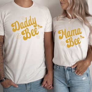 May include: Two white t-shirts with the text "Daddy Bee" and "Mama Bee" in yellow script. Each shirt features a small bee graphic and honeycomb designs.
