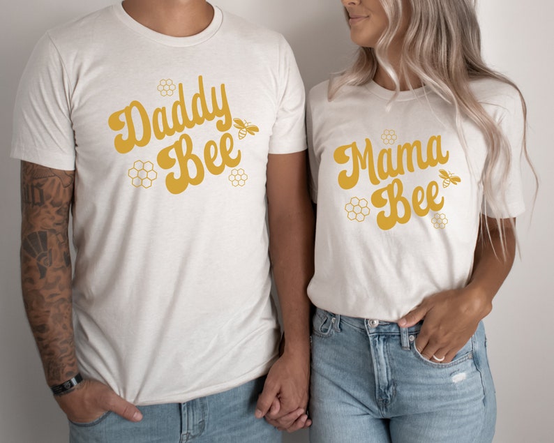 May include: Two white t-shirts with the text "Daddy Bee" and "Mama Bee" in yellow script. Each shirt features a small bee graphic and honeycomb designs.