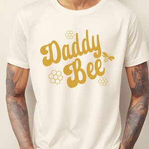May include: White t-shirt with a gold graphic that says "Daddy Bee" with a stylized bee illustration.