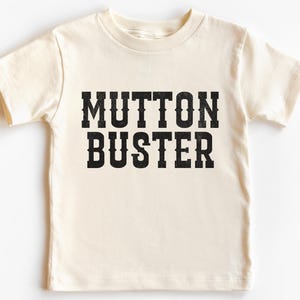 May include: Cream-colored t-shirt with the words "MUTTON BUSTER" printed in bold, black, western-style lettering. The shirt has short sleeves and a crew neck.