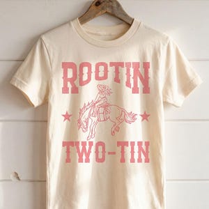 May include: A cream-colored short-sleeve t-shirt featuring a pink graphic print. The design includes the text ROOTIN TWO-TIN in a western-style font, with a central illustration of a person riding a bucking horse, flanked by two pink stars.