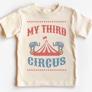 May include: A cream-colored t-shirt with a blue and red circus tent graphic and the text "My Third Circus" in blue and red.