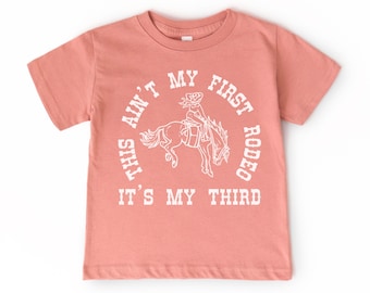 May include: A pink t-shirt with a white graphic that says "This ain't my first rodeo, it's my third". The graphic also includes a drawing of a cowgirl riding a horse.