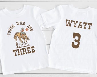 May include: Two white t-shirts, one with a cowboy riding a horse and the text "Young, Wild, and Three", the other with the name "Wyatt" and the number "3" in brown text.