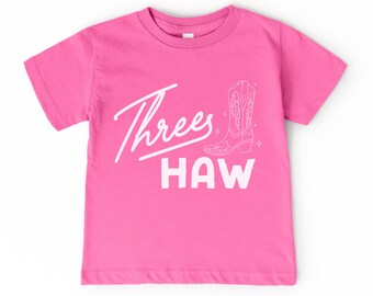 May include: A bright pink t-shirt with the words "Threes Haw" in white script and block letters. A white outline of a cowboy boot is also featured. The shirt is a short-sleeved crew neck.