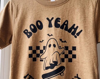 May include: A tan short-sleeve t-shirt featuring a black graphic print of a ghost on a skateboard, a checkered pattern, and three small bats. The text reads BOO YEAH! IT'S MY BIRTHDAY in a bold, curved font centered on the chest of the shirt.