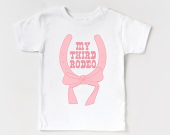May include: White t-shirt with a pink horseshoe and bow design. The text "MY THIRD RODEO" is printed on the shirt.