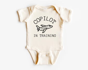 Peut inclure: Body de bébé crème avec l'inscription "COPILOT" au-dessus d'un dessin d'avion noir et les mots "IN TRAINING" en dessous. Le body a des manches courtes et une fermeture à boutons-pression.