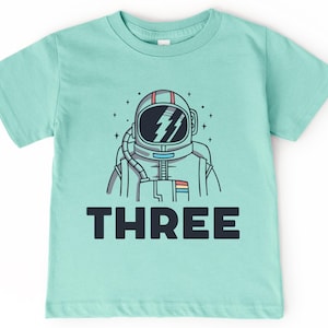 May include: A light blue t-shirt with a cartoon astronaut wearing a helmet and the word "THREE" printed on the front.