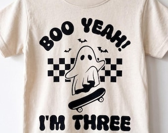 May include: A cream-colored short-sleeve t-shirt featuring a black graphic of a ghost on a skateboard, a checkered pattern, three small bats, and the text "BOO YEAH! I'M THREE" printed in a bold, rounded black font on the front of the garment.