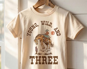 May include: A cream-colored short-sleeve t-shirt featuring a brown graphic print of a person riding a bucking horse. The text reads YOUNG, WILD, AND THREE. The shirt is displayed on a wooden hanger against a white horizontal paneled wall.