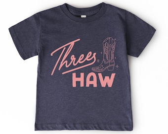 May include: Navy blue t-shirt with a pink graphic that says "Threes Haw" and a drawing of a cowboy boot. The shirt is made of a soft material and is suitable for a child.