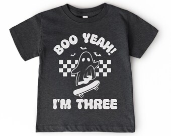May include: Dark gray t-shirt with a white graphic of a ghost on a skateboard, the text "BOO YEAH!" above, and "I'M THREE" below. The design includes bats and a checkered pattern, perfect for a Halloween or birthday theme.